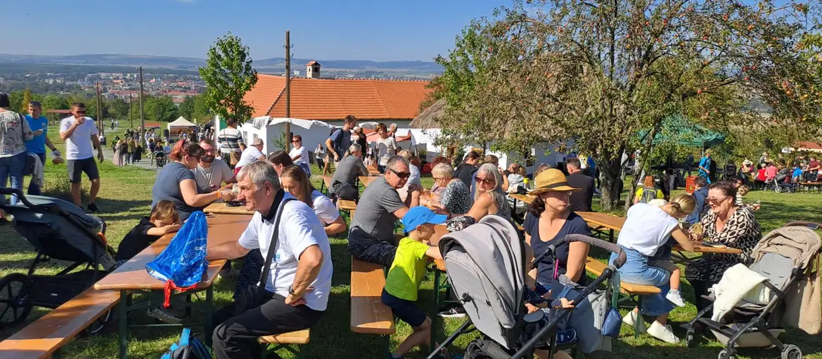 Plné lavice a stoly pod stromy na Slováckém festivalu chutí a vůní, v pozadí výhled na Uherské Hradiště
